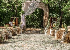 La imagen presenta un photocall de boda con un estilo rústico, compuesto de un marco a medida, y que imita a un paraje de la toscana italiana, adornado con una cortina y flores diversas como geranios y girasoles.