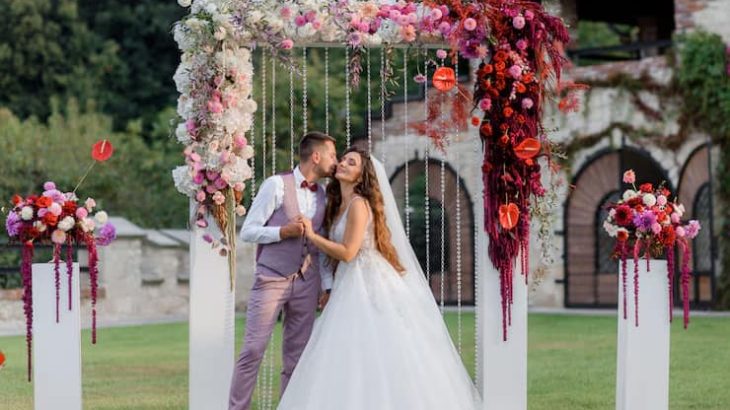 En la imagen se muestra a una pareja que acaba de casarse, haciéndose una foto en un photocall elegante que se ha instalado en su boda. El photocall se compone de un marco a medida blanco adornado con flores muy variadas.