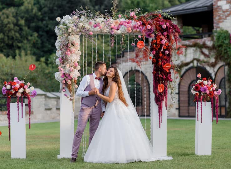En la imagen se muestra a una pareja que acaba de casarse, haciéndose una foto en un photocall elegante que se ha instalado en su boda. El photocall se compone de un marco a medida blanco adornado con flores muy variadas.