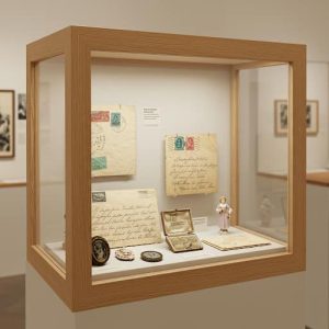 En la foto se pesenta una vitrina dentro de lo que parece un museo, hecha a medida a partir de un cartón pluma, marcos y metacrilato, dentro de la cual se hallan objetos de valor como cartas, monedas, figuras y algún que otro sello
